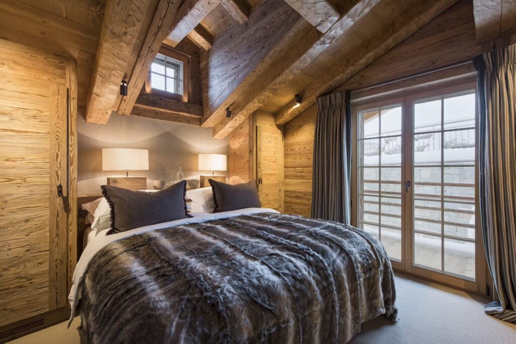 A cozy bedroom with wood-paneled walls and ceiling, a large bed with fur blanket and pillows, two bedside lamps, and glass doors opening to a balcony with a snowy view. David Pearson Travel