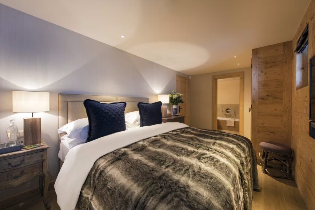 A cozy bedroom with a double bed covered in a plush fur blanket, two navy blue pillows, wooden furniture, soft lighting from bedside lamps, and an open door revealing a bathroom. David Pearson Travel