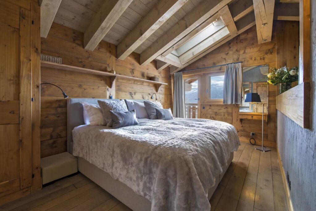 A cozy bedroom with wooden walls and ceiling, a large bed with gray bedding, shelves, a skylight, and a window with curtains letting in natural light. There’s a small desk, chair, and a vase of flowers in the corner. David Pearson Travel