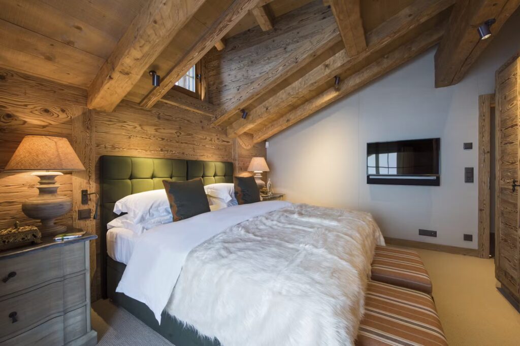 Cozy bedroom with rustic wooden walls and ceiling beams, a large bed with white bedding and green headboard, two bedside tables with lamps, a wall-mounted TV, and soft lighting creating a warm atmosphere. David Pearson Travel