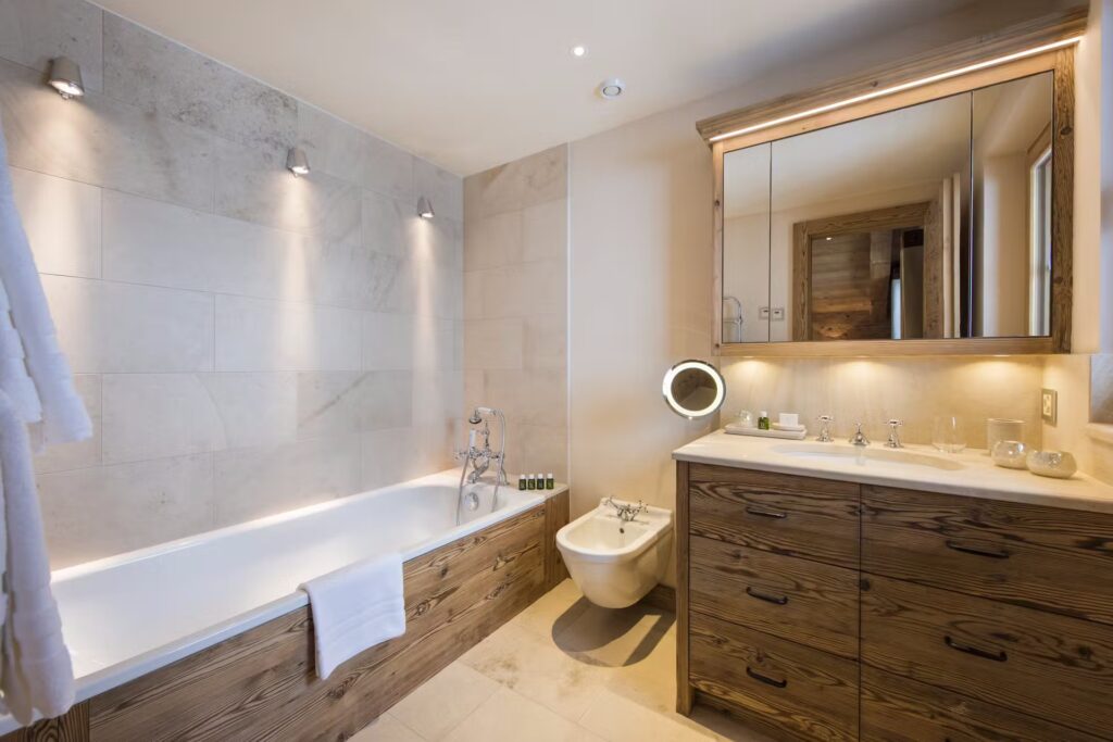 Modern bathroom with a built-in bathtub, bidet, and wooden vanity with a large mirror; soft lighting and beige tiles create a warm, inviting atmosphere. A towel hangs on the edge of the tub. David Pearson Travel