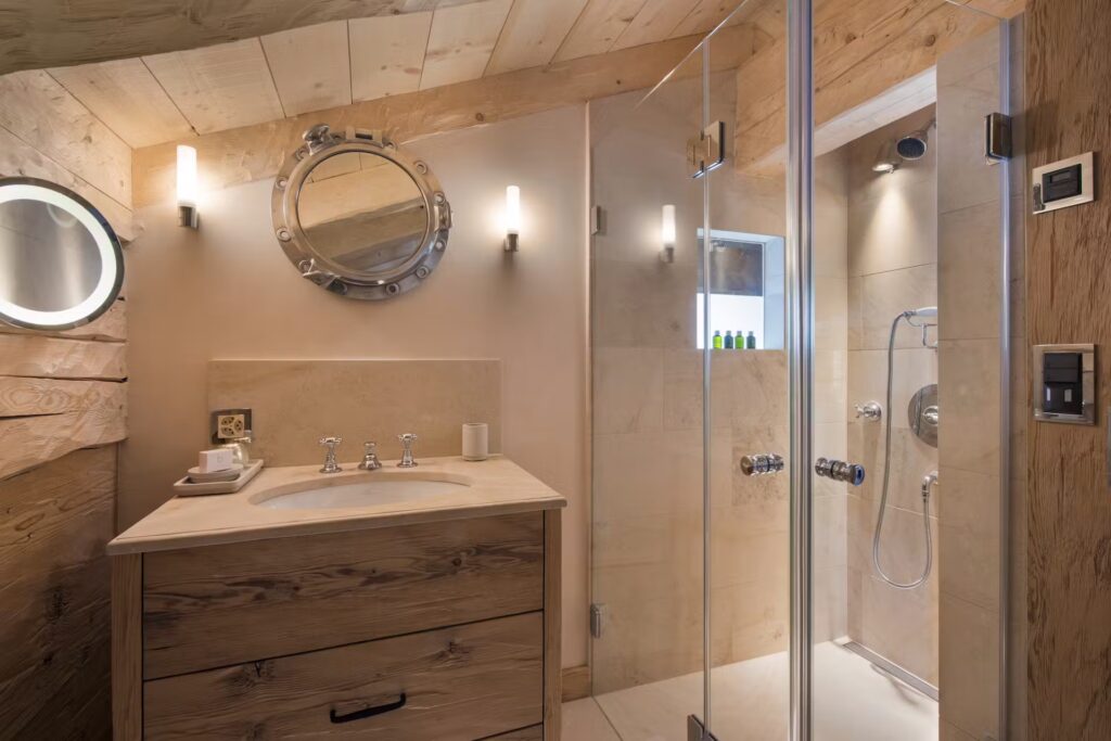 Modern bathroom with wooden accents, round mirrors, wall-mounted lights, a glass shower enclosure, and a sink with a wooden vanity. Neutral tones and natural materials create a warm, inviting atmosphere. David Pearson Travel