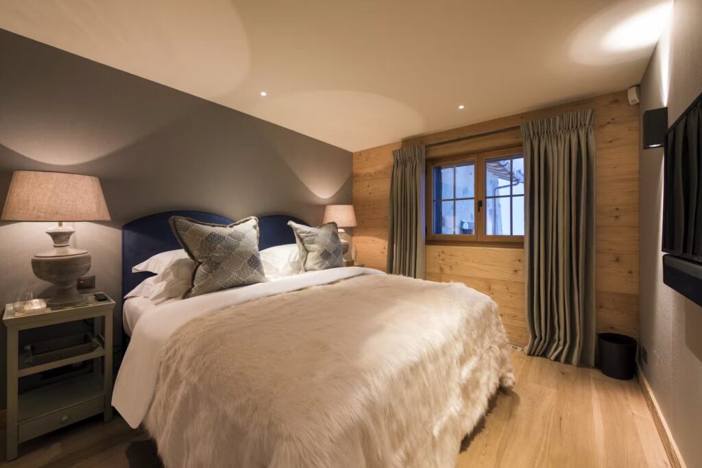 A cozy bedroom with a large bed covered in a furry beige blanket, decorative pillows, two bedside tables with lamps, wooden walls, a window with curtains, and a wall-mounted TV. Warm lighting creates a relaxing atmosphere. David Pearson Travel