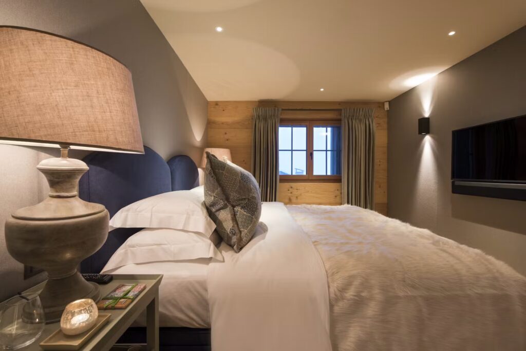 A cozy, warmly lit bedroom with a large bed, fur blanket, decorative pillows, bedside lamp, and a tray with candles. There is a window with curtains at the end of the room and a wall-mounted TV. David Pearson Travel