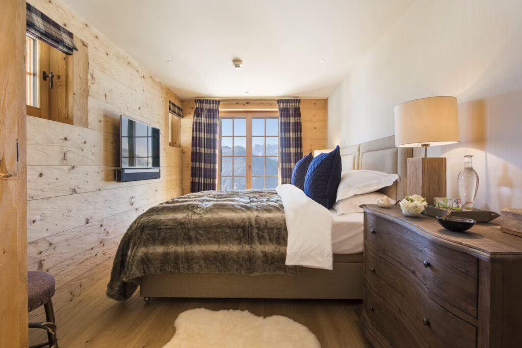 Cozy bedroom with wooden walls, a large bed covered in a fur blanket, blue accent pillows, a wall-mounted TV, and a window with plaid curtains letting in natural light. A wooden dresser with a lamp and decor sits beside the bed. David Pearson Travel