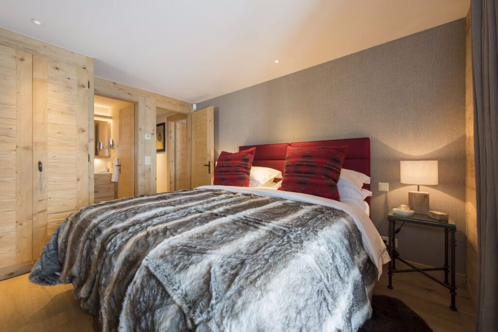 A cozy bedroom with a large bed covered in a faux fur blanket, two red pillows, and a bedside table with a lamp. Wooden doors and walls add a warm, rustic touch; a bathroom is visible through an open door. David Pearson Travel