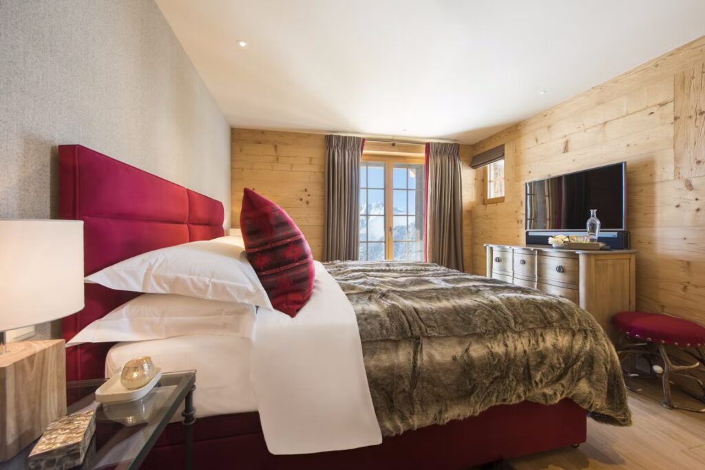 A cozy bedroom with wood-paneled walls, a large bed with a red headboard and fur blanket, a glass bedside table with a lamp, a dresser with a TV, and a window letting in natural light. David Pearson Travel