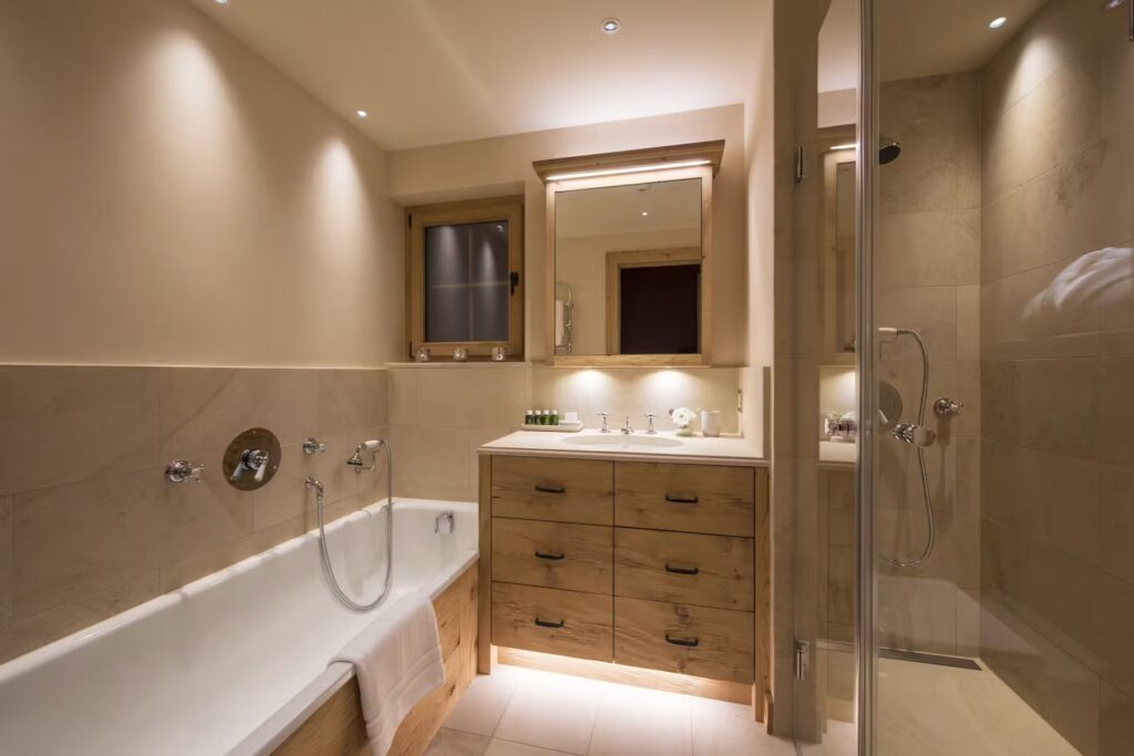 A modern bathroom with a bathtub, a wooden vanity with a large mirror, a shower with glass doors, beige tiled walls, and soft recessed lighting. David Pearson Travel
