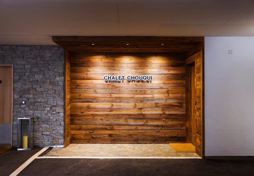 A wooden entryway with the sign CHALET CHOUQUI mounted on horizontal wood panels, next to a stone wall and a glass door; warm ceiling lights illuminate the area. David Pearson Travel