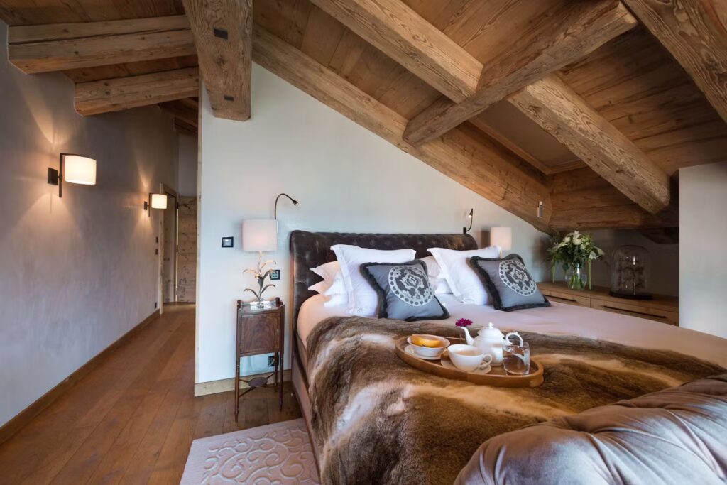 A cozy bedroom with a sloped wooden ceiling, a large bed covered with pillows and a fur throw, a tray with breakfast on the bed, bedside tables, wall lights, and a hallway with wooden floors. David Pearson Travel