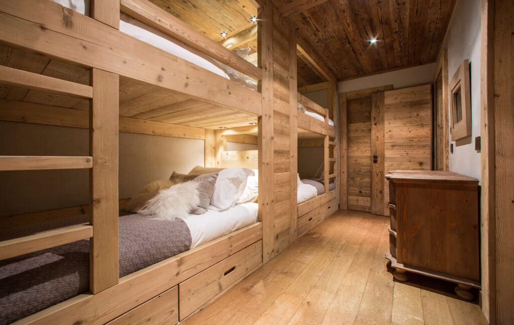 A cozy bedroom with wooden walls, ceiling, and floor featuring two sets of bunk beds, a small wooden dresser, and warm lighting, creating a rustic, cabin-like atmosphere. David Pearson Travel
