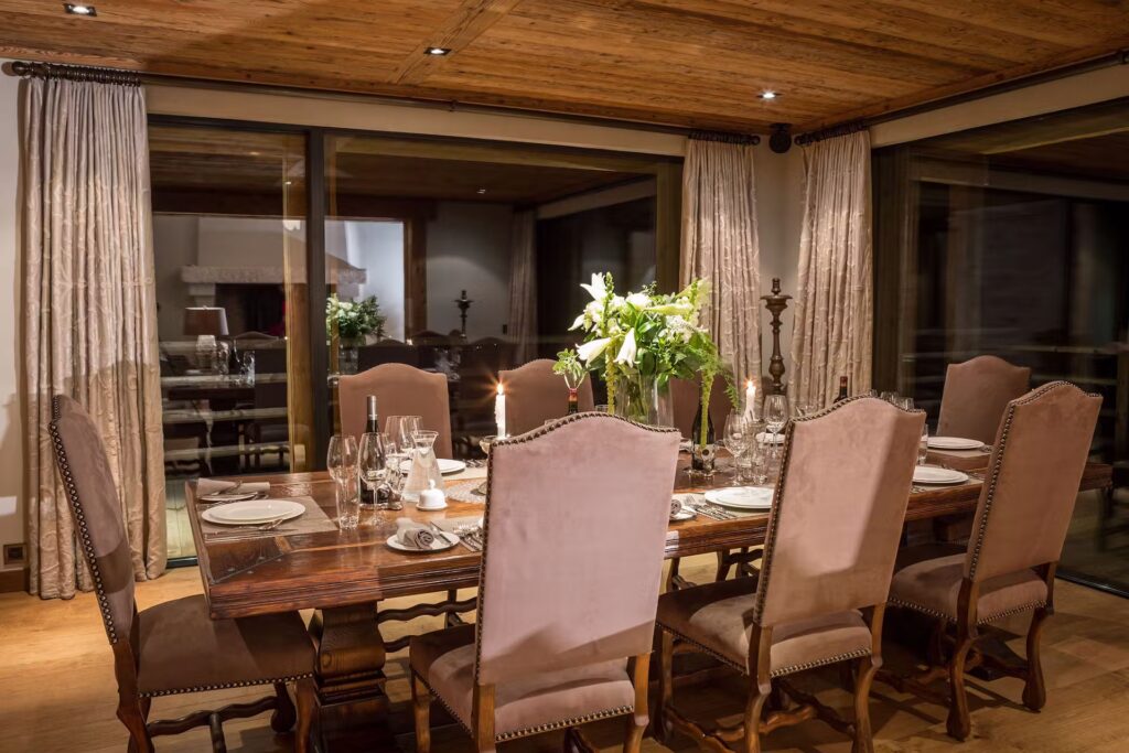 A warmly lit dining room with a wooden table set for eight, surrounded by high-back upholstered chairs. The table features elegant dinnerware, glassware, candles, and two vases of white flowers. Large curtains frame the windows. David Pearson Travel