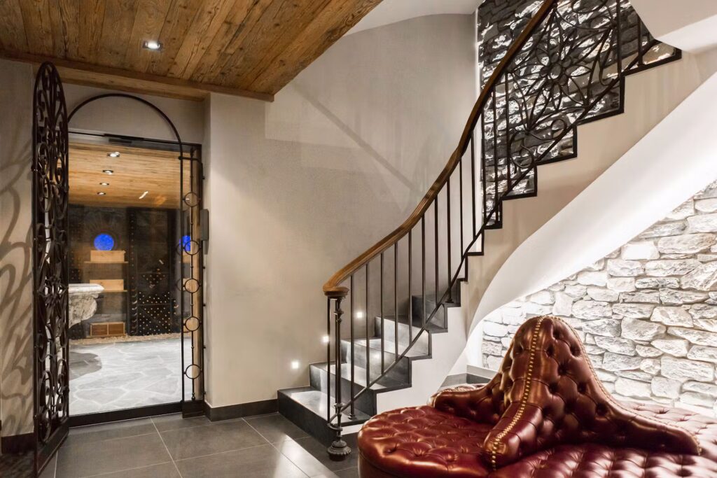 A modern entryway with a curved staircase featuring a black ornate railing, a plush red tufted chair, stone accent wall, wooden ceiling, and a glass door leading to a wine cellar with stone flooring and wine storage racks. David Pearson Travel