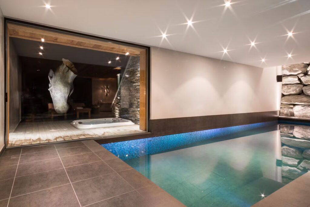 Modern indoor pool with clear water, gray tiled floor, and a large glass wall revealing a hot tub and rustic stone decor in an adjacent room. Ceiling spotlights create a bright, inviting atmosphere. David Pearson Travel