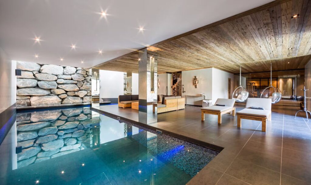 Luxurious indoor pool area with stone accent wall, wooden ceiling, modern lounge chairs, and a seating area, all in a spacious, well-lit room with contemporary decor. David Pearson Travel