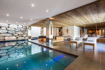 Luxurious indoor pool area with stone accent wall, wooden ceiling, modern lounge chairs, and a seating area, all in a spacious, well-lit room with contemporary decor. David Pearson Travel
