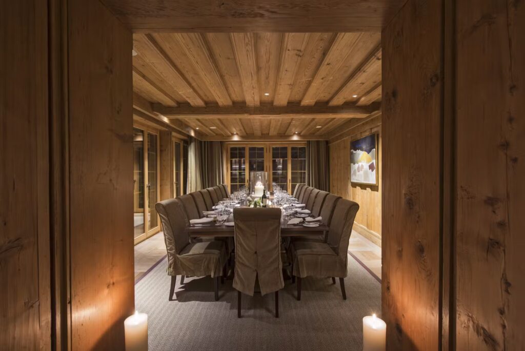 A warmly lit dining room with wood-paneled walls and ceiling, a long table set for a meal with elegant place settings, brown upholstered chairs, candles, and large windows along the side. David Pearson Travel
