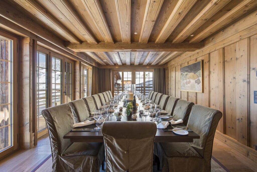A rustic dining room with a long wooden table set for a meal, surrounded by cushioned chairs. The room features wooden walls, a beamed ceiling, large windows, and mountain views. David Pearson Travel