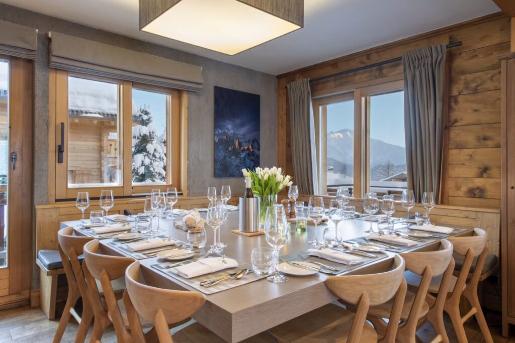 Elegant dining room with wooden walls and large windows showing snowy scenery. A square table is set for twelve with wine glasses, plates, napkins, and a vase of white tulips in the center. Natural light fills the space. David Pearson Travel