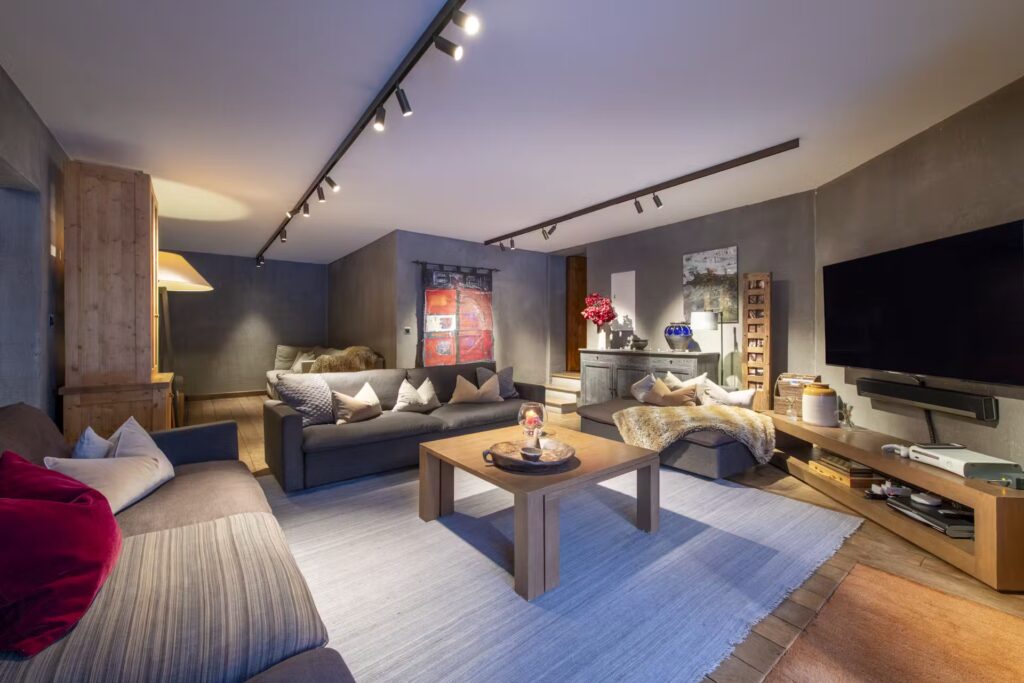 Modern living room with two sofas, a wooden coffee table, a large TV on a wooden stand, track lighting on the ceiling, decorative pillows and art, and a cozy, inviting atmosphere with neutral tones and warm accents. David Pearson Travel