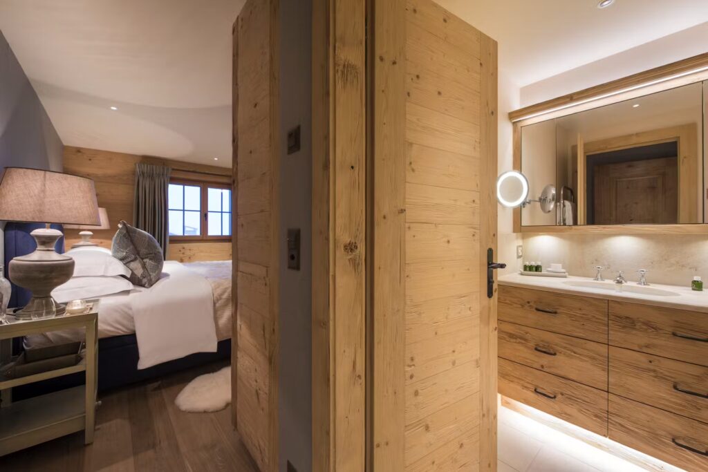 A cozy bedroom and modern bathroom separated by a wooden wall; the bedroom has a bed with white linens and a bedside lamp, while the bathroom features a wide mirror, sink, and wooden cabinets. David Pearson Travel