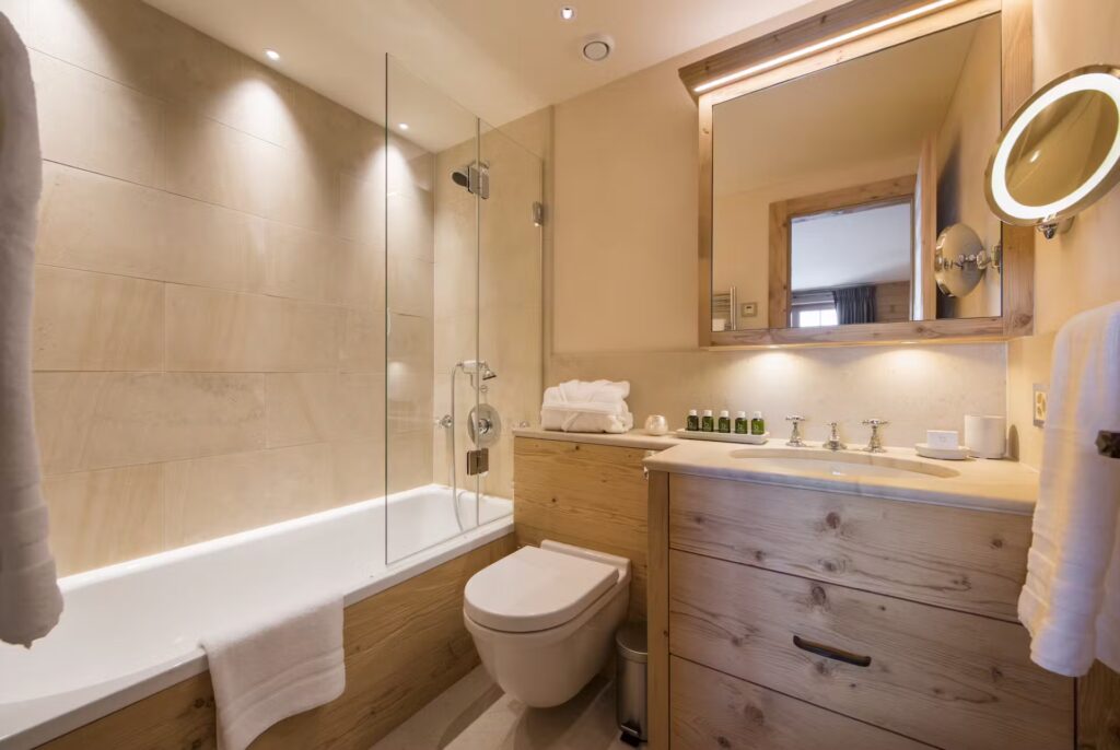 Modern bathroom with a glass-enclosed bathtub, wall-mounted toilet, wooden vanity, large mirror, towel rack, and neatly arranged toiletries on the countertop under soft overhead lighting. David Pearson Travel