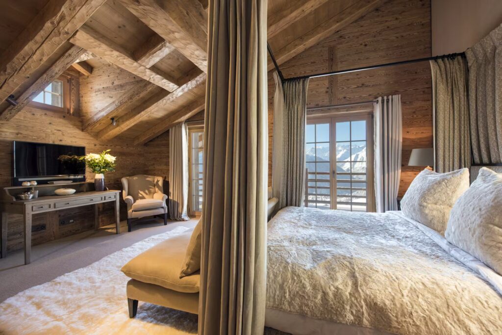 Cozy attic bedroom with wooden beams, a large bed with fur bedding, light curtains, an armchair, TV on a wooden console, and floor-to-ceiling windows revealing mountain views. David Pearson Travel