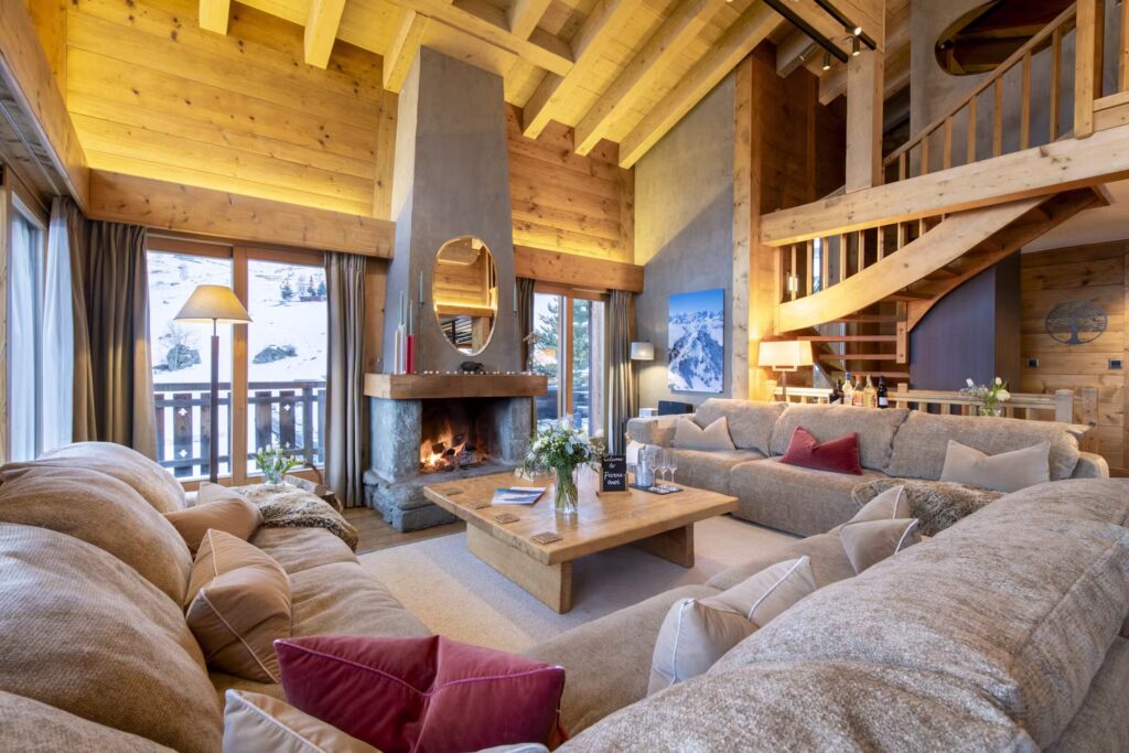 A cozy, modern alpine living room with wooden walls and ceiling, large windows, a stone fireplace, beige sofas with colorful cushions, a wooden coffee table, and a spiral staircase leading to an upper level. David Pearson Travel