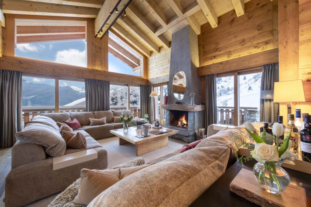 A cozy, modern chalet living room with large windows showing snowy mountain views, plush sofas, a stone fireplace, wooden beams, and warm lighting. Flowers and decor items sit on tables, creating an inviting atmosphere. David Pearson Travel