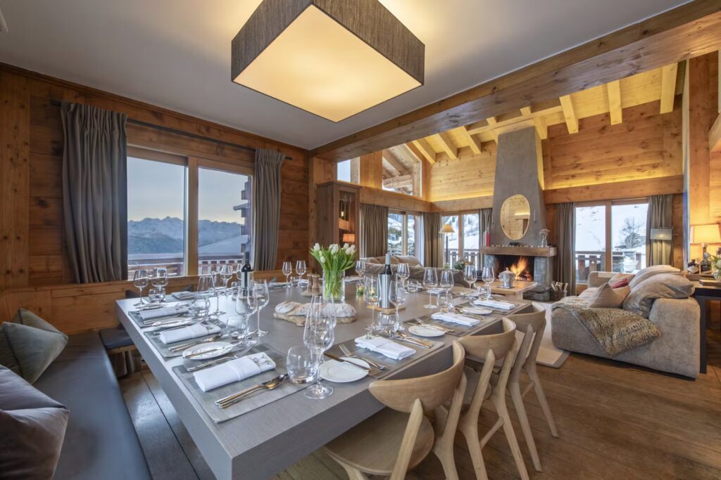 A cozy, modern dining room with a large wooden table set for twelve, surrounded by light wooden chairs. A fireplace, plush seating, wood-paneled walls, and large windows offer mountain and forest views. David Pearson Travel