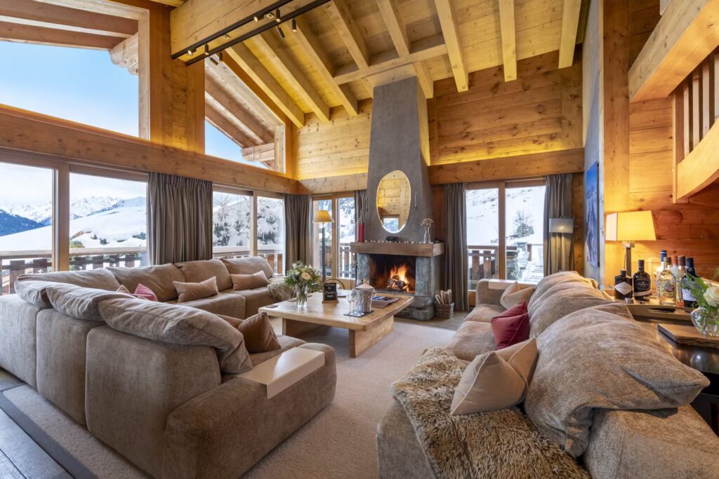A cozy, spacious chalet living room with large windows overlooking snowy mountains. There are plush sofas, a central coffee table, a roaring fireplace, wooden beams, and warm ambient lighting. David Pearson Travel
