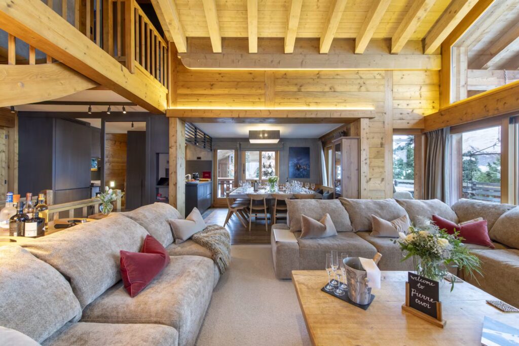 Spacious, cozy living room with beige sofas, red cushions, wooden beams, a dining area with a long table, and large windows letting in natural light. A bar cart and flower arrangement are visible. David Pearson Travel