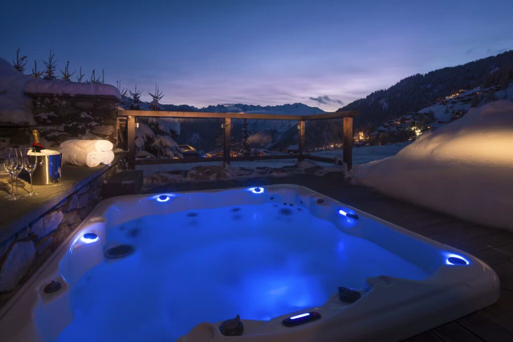 A glowing outdoor hot tub with blue lights sits on a snowy deck at dusk, overlooking a mountain village. Rolled towels, champagne, and glasses are set nearby, creating a cozy, inviting winter scene. David Pearson Travel