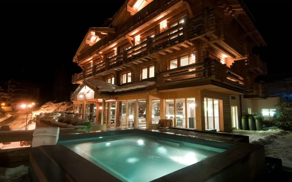 A warmly lit wooden chalet at night with large windows, snowy surroundings, and a glowing outdoor hot tub in the foreground. David Pearson Travel