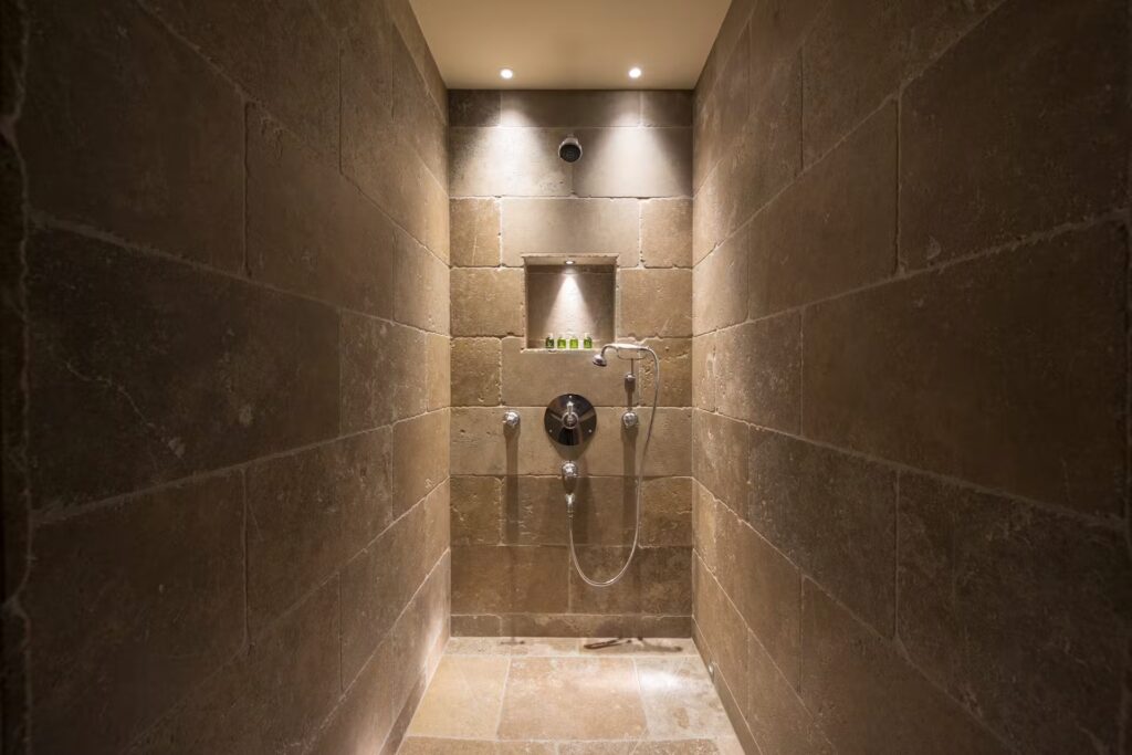 A modern walk-in shower with brown stone walls, built-in shelf holding small toiletry bottles, rainfall showerhead, and recessed ceiling lights creating a warm, inviting atmosphere. David Pearson Travel