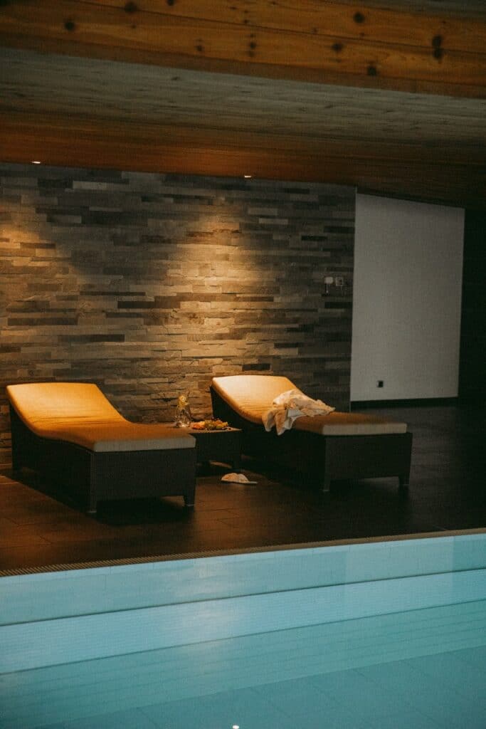 Two lounge chairs with cushions sit beside an indoor pool, against a stone accent wall and under a wooden ceiling, with soft lighting creating a relaxing atmosphere. A towel and drink are on the chairs. David Pearson Travel
