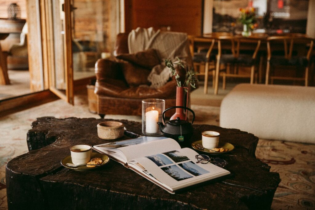 A cozy living room with an open book, two cups of coffee, cookies, a lit candle, and a teapot on a rustic wooden table. A leather armchair and dining area are visible in the warm, softly lit background. David Pearson Travel