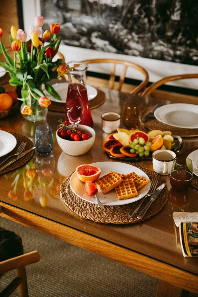 A breakfast table set with waffles, sliced grapefruit, fruit, coffee, a pitcher of juice, and a bowl of strawberries. Colorful tulips and plates are also on the wooden table. David Pearson Travel