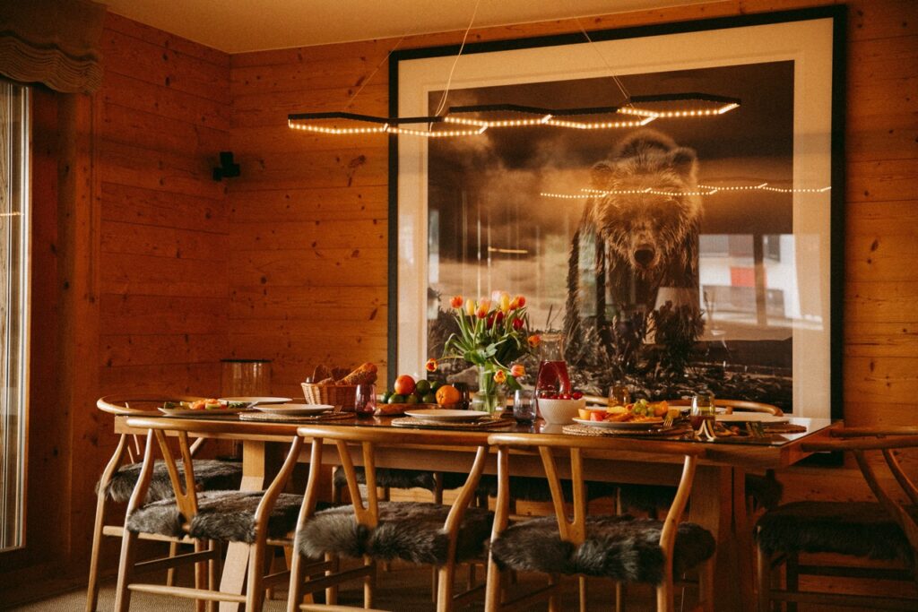 A cozy dining room with wooden walls, a long table set for a meal, fur-covered chairs, fresh flowers, and a large framed bear photograph on the wall behind the table. Warm lighting creates an inviting atmosphere. David Pearson Travel