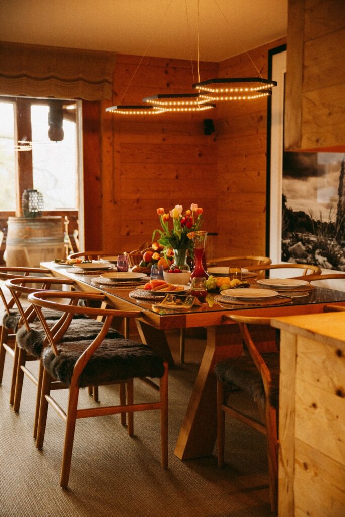 A cozy wooden dining room with a set table featuring tulips in a vase, various fruits, glassware, placemats, and fur-covered chairs, illuminated by warm hanging lights. David Pearson Travel