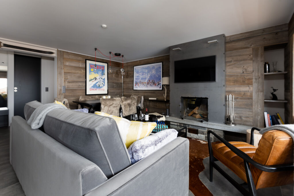 A modern living room with wood-paneled walls, a gray sofa, a brown leather chair, a wall-mounted TV above a fireplace, and framed artwork. Shelves, books, and decorative items are placed around the room. David Pearson Travel