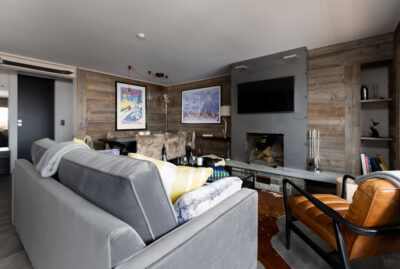 A cozy living room with a gray sofa, brown leather chair, wooden coffee table, and modern fireplace evokes the inviting charm found in luxury Verbier ski chalet rentals—art, blankets, and rich decor create a warm, welcoming atmosphere. David Pearson Travel