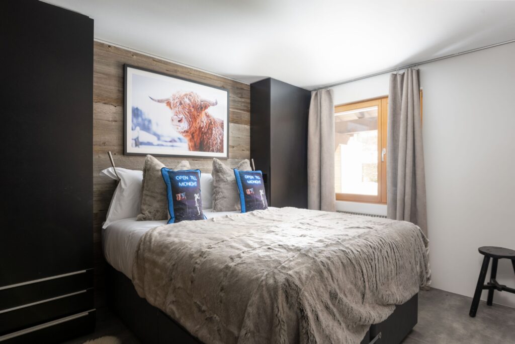 A cozy bedroom with a plush bed covered in a soft gray blanket, decorative pillows, a wooden accent wall, framed cow art above the bed, two curtains framing a window, and a black stool in the corner. David Pearson Travel
