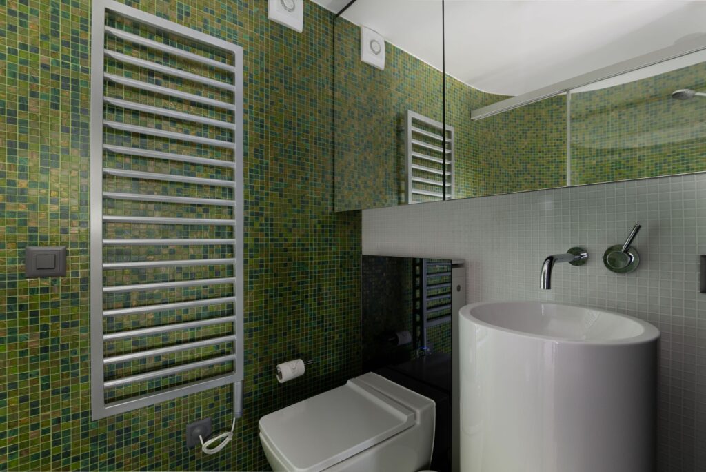 Modern bathroom with green mosaic tile walls, a wall-mounted toilet, a cylindrical white sink, a metallic towel radiator, a toilet paper holder, and a mirrored upper wall reflecting the room. David Pearson Travel