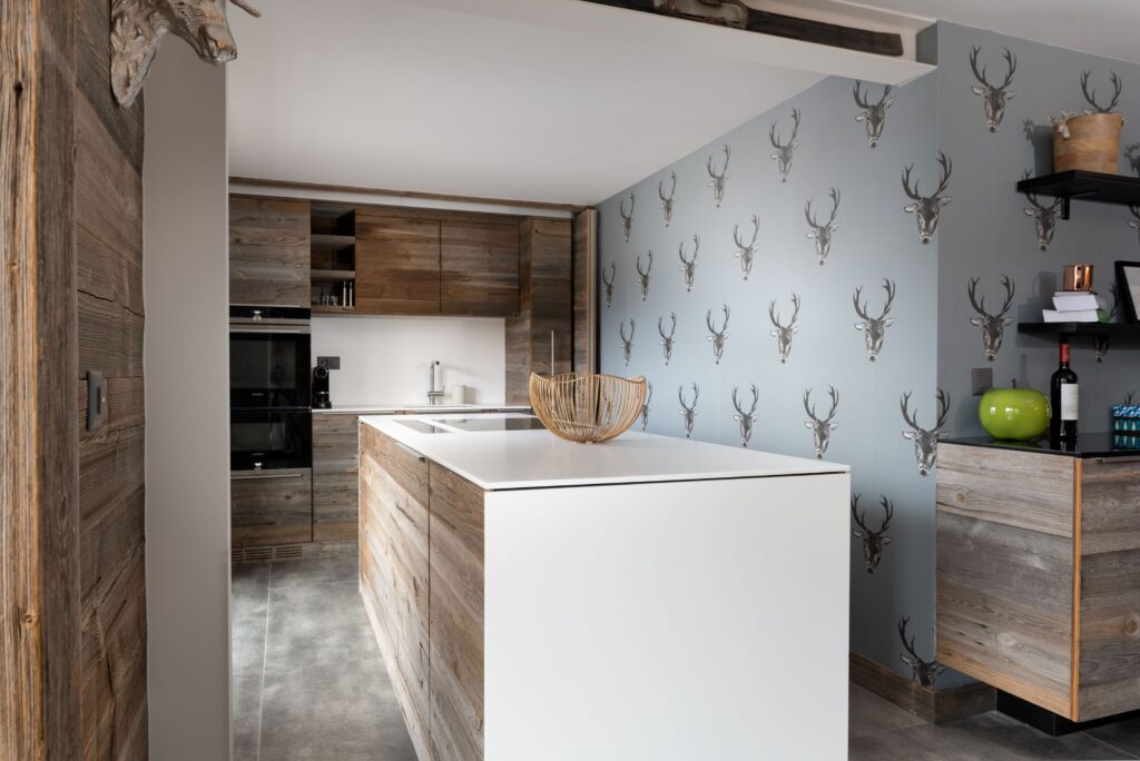 Modern kitchen with rustic wooden cabinets and island, minimalist white countertops, and a gray wall decorated with a repeating deer head pattern. A wicker basket sits on the island; shelves hold kitchen items. David Pearson Travel