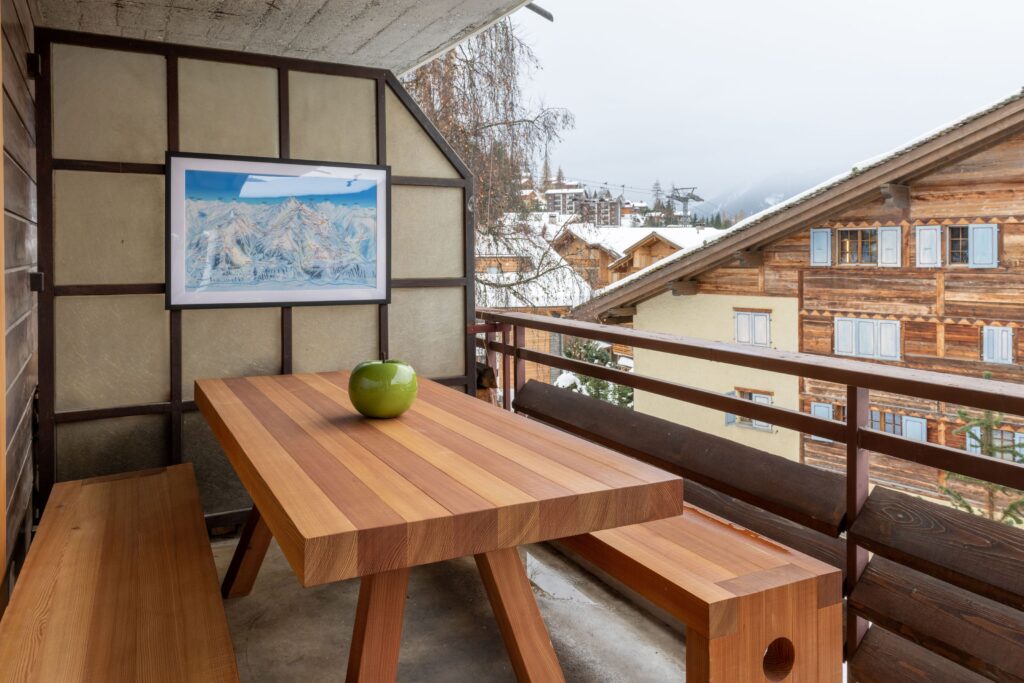 A wooden table with matching benches on a balcony, holding a single green apple in a bowl. A framed mountain painting hangs on the wall, with snowy chalet-style buildings visible outside. David Pearson Travel