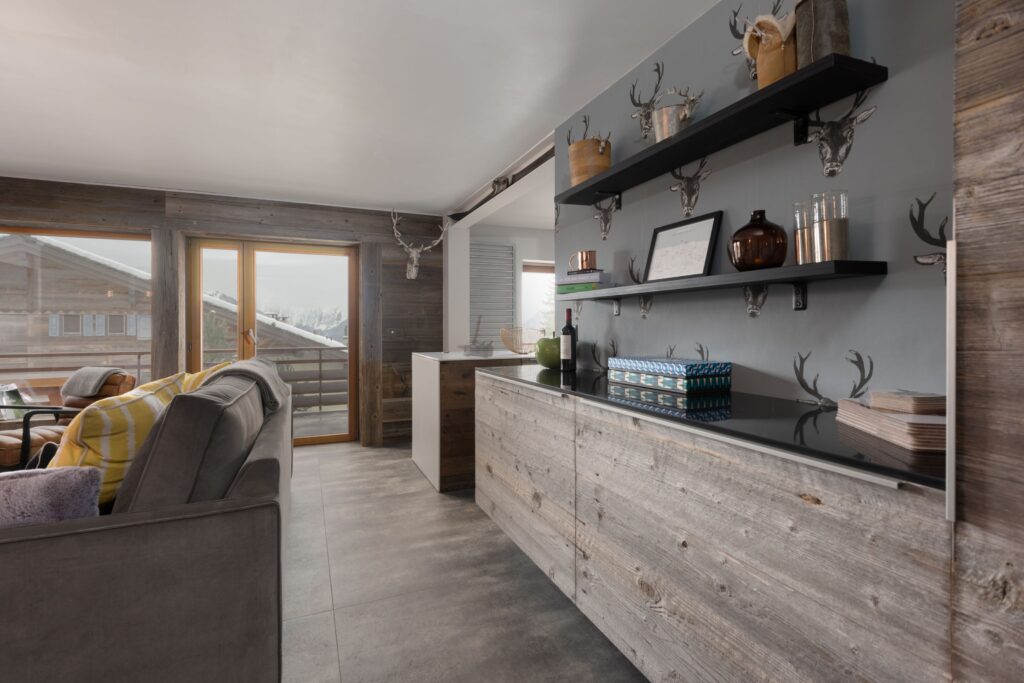 Modern rustic living space with gray wooden cabinets, floating shelves with decorative items, a gray sofa with yellow plaid blanket, large windows, and mountain views outside. Neutral tones and natural light fill the room. David Pearson Travel