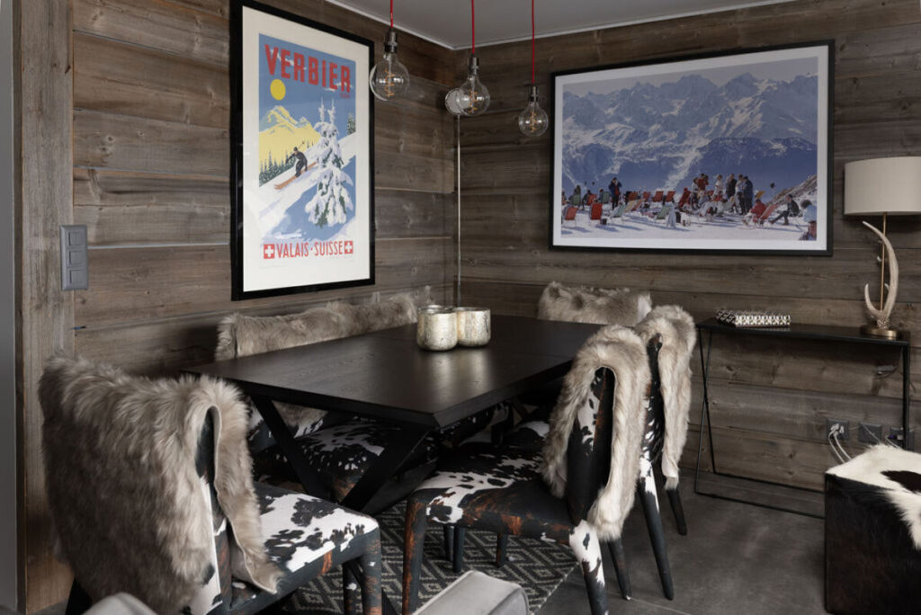 Cozy dining area with a dark wooden table, fur-draped and cowhide-patterned chairs, rustic wood walls, two framed ski posters, and glass pendant lights hanging above the table. David Pearson Travel