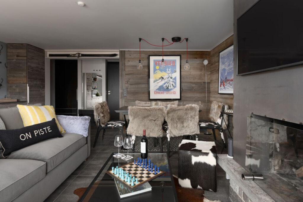 Modern living room with a gray sofa, fur-covered dining chairs, rustic wood walls, a wall-mounted TV, a fireplace, alpine posters, and a glass coffee table with a chess set, wine glasses, and a On Piste pillow. David Pearson Travel