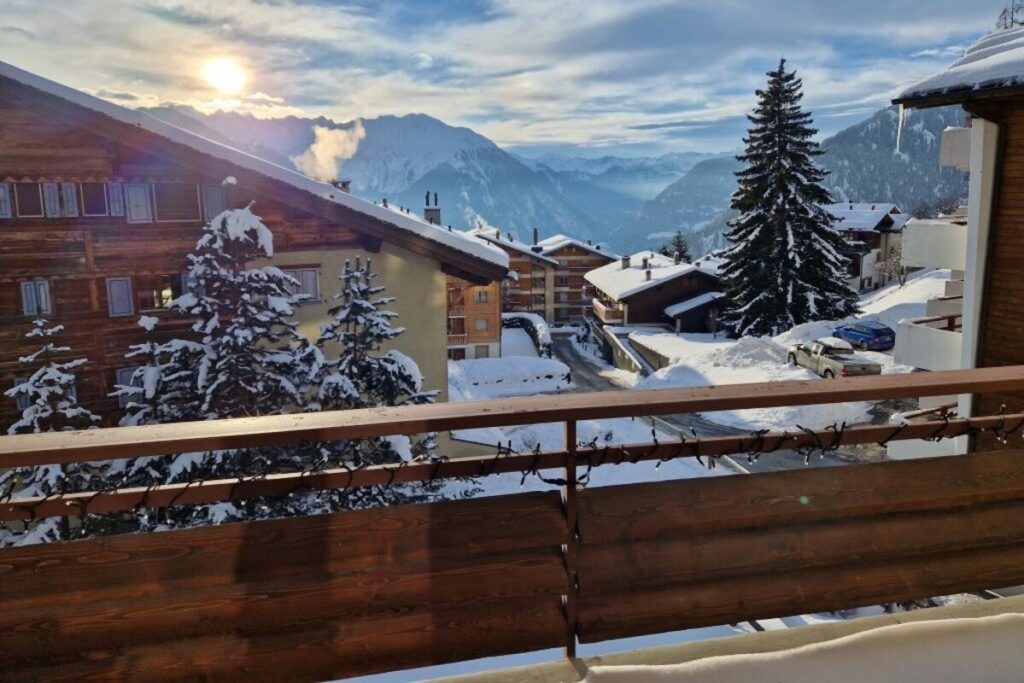 View from a wooden balcony overlooking snow-covered chalets, pine trees, and mountains in the background under a partly cloudy sky with the sun shining. David Pearson Travel